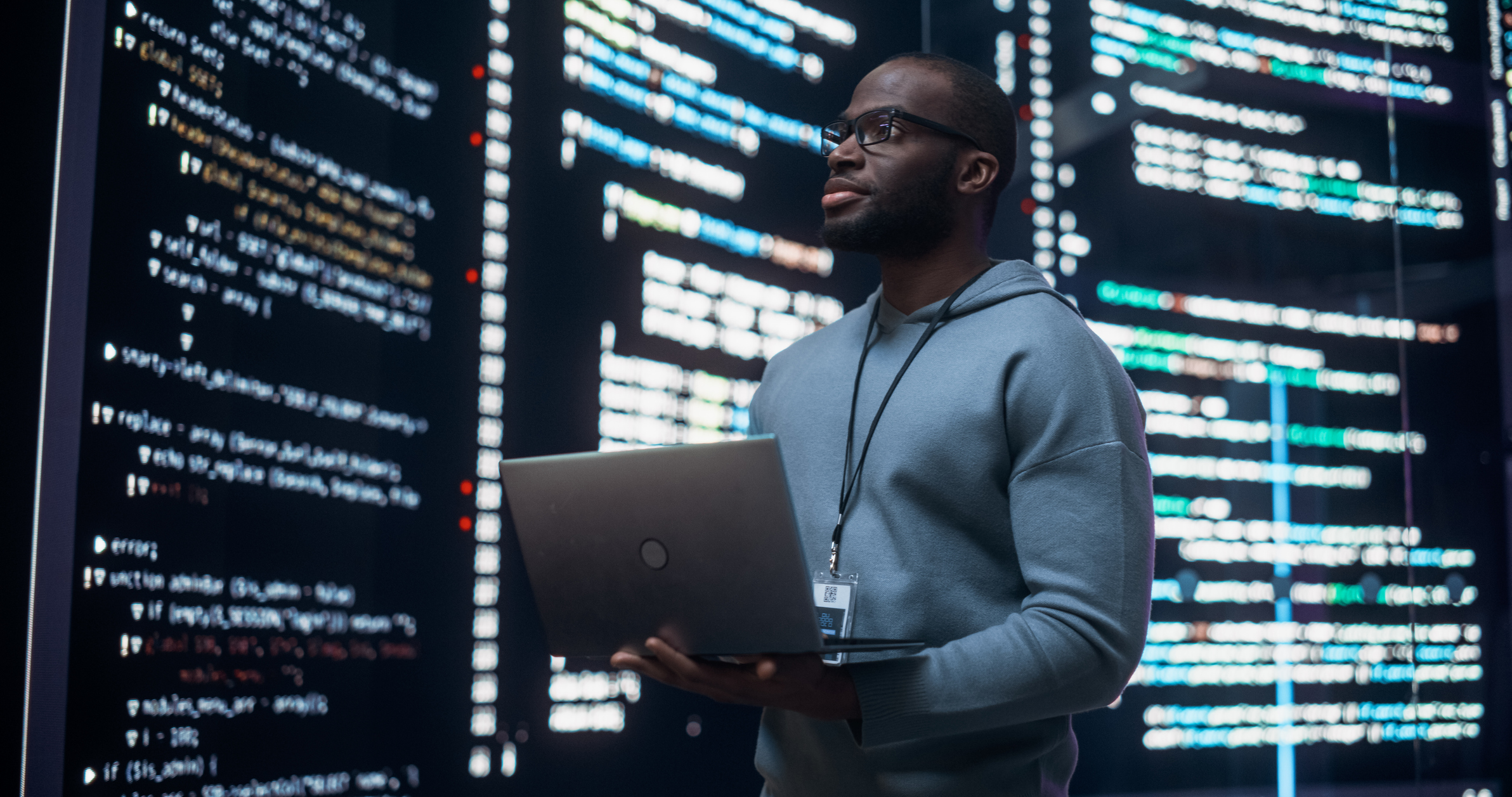A man standing in front of a series of screens holding a laptop while reviewing data